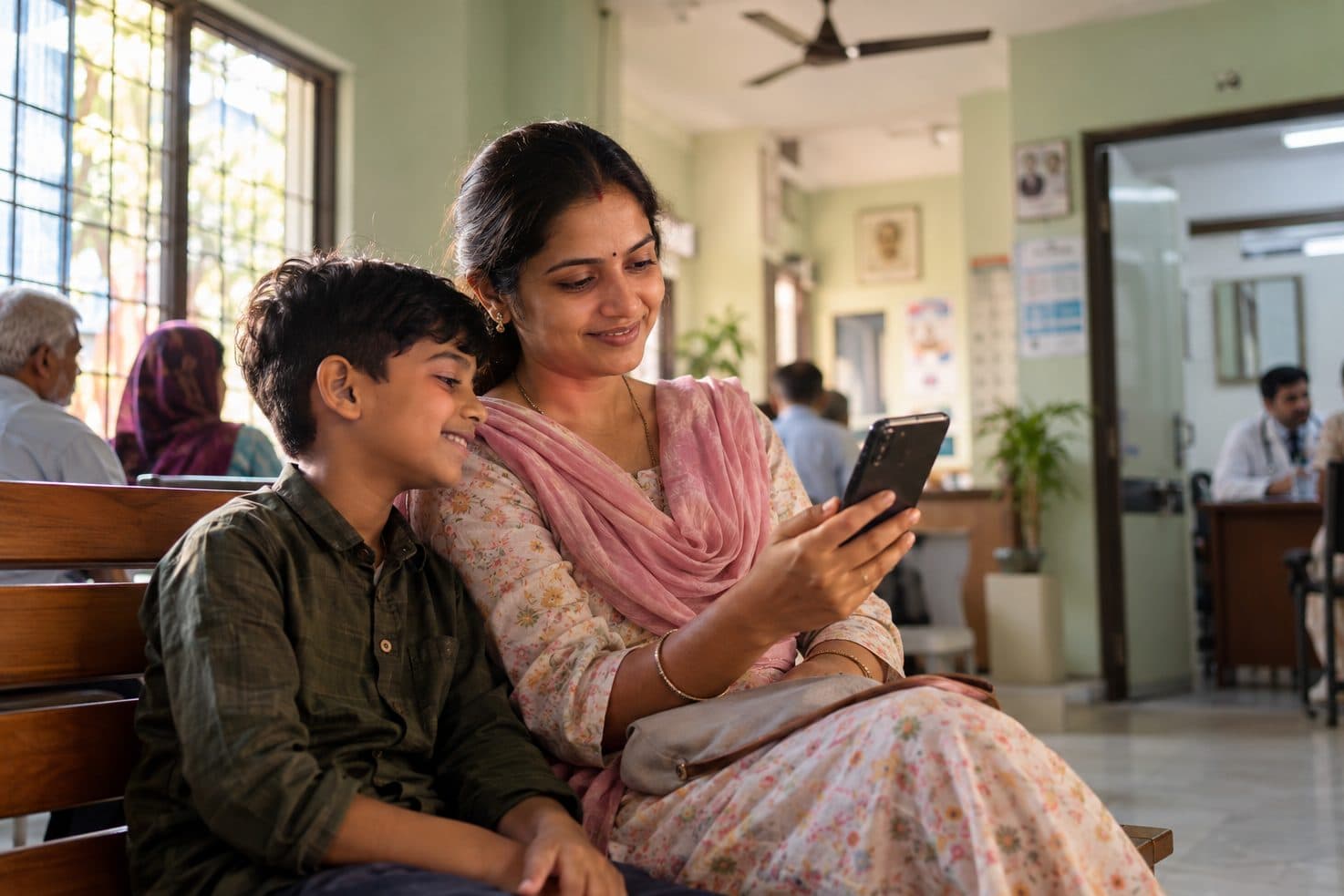ક્લિનિકના વેઇટિંગ રૂમમાં ફોન જોઈને હસતાં માતા અને પુત્ર